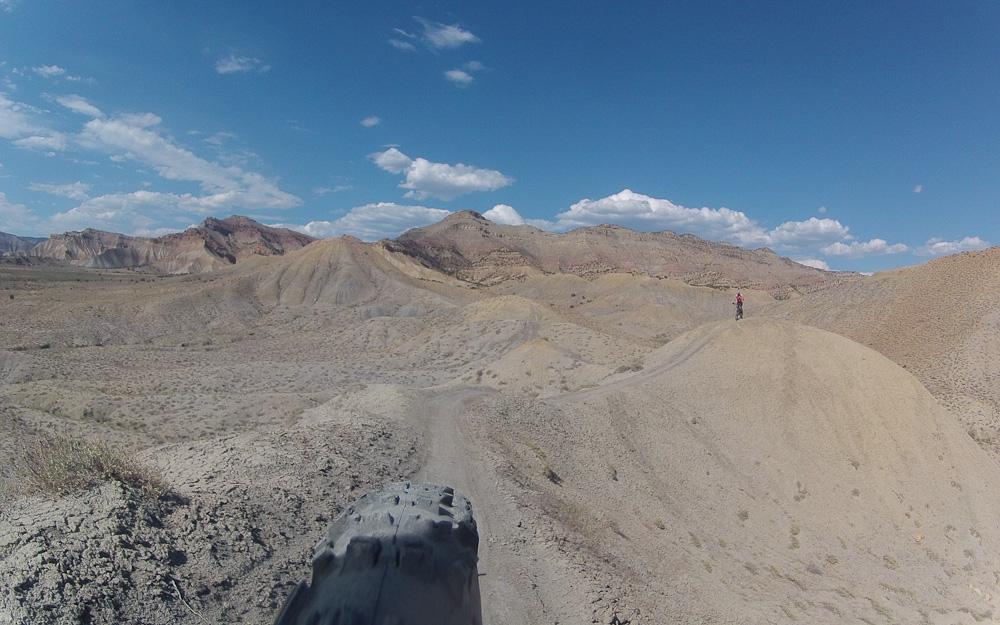 A scenic view of a rugged, desolate landscape featuring rolling hills and dry terrain, with a mountain biker in a red shirt standing on a hill in the distance. The sky is blue with scattered clouds, and the foreground includes a close-up of a mountain bike tire. Zippety Do Dah mountain bike trail.