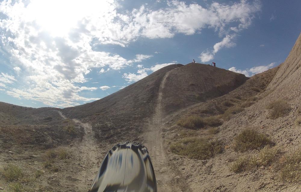 A view of a dirt bike trail leading up to a hill, with a clear blue sky and scattered clouds overhead. The foreground shows part of a bike tire, while two cyclists are seen at the top of the hill in the background. The landscape features dry, rugged terrain with sparse vegetation. Zippety Do Dah mountain bike trail.