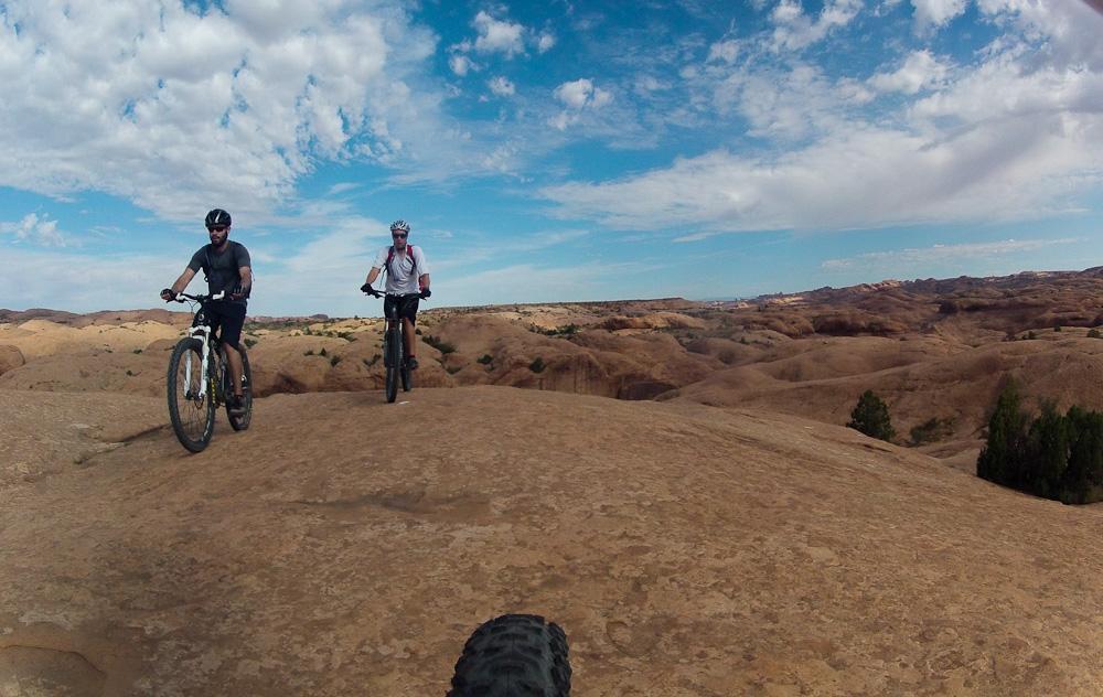 Two mountain bikers riding on a rocky terrain with a backdrop of expansive desert landscape and blue sky dotted with clouds. The first rider, wearing a helmet and dark clothing, is in the foreground, while the second rider, dressed in lighter gear, follows closely behind. Slickrock mountain bike trail.