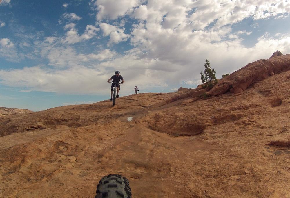 Two mountain bikers navigating a rocky terrain under a partly cloudy sky. The foreground shows a close-up of a bike tire, while one biker ascends a slope ahead. The landscape features reddish rock formations and sparse vegetation. Slickrock mountain bike trail.