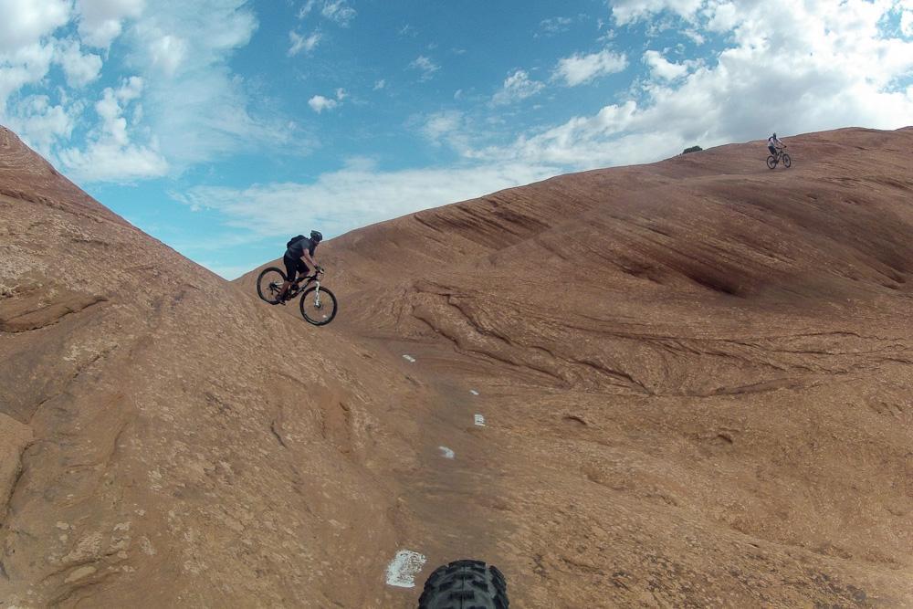 A mountain biker navigating a rocky terrain against a backdrop of blue sky and scattered clouds. The cyclist is descending a steep, uneven surface, while another biker is visible in the background on a higher elevation. Slickrock mountain bike trail.