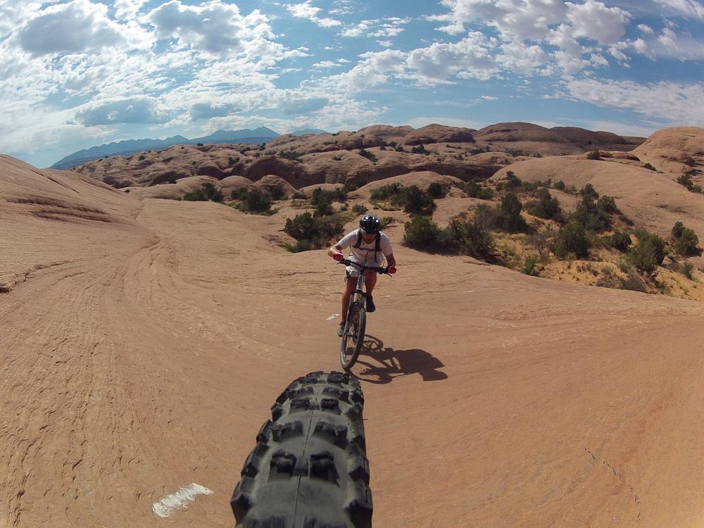 A cyclist riding a mountain bike on a rocky terrain under a partly cloudy sky, with hills and vegetation in the background. The photo captures the perspective of the bicycle's front tire aiming forward as the rider navigates the landscape. Slickrock mountain bike trail.