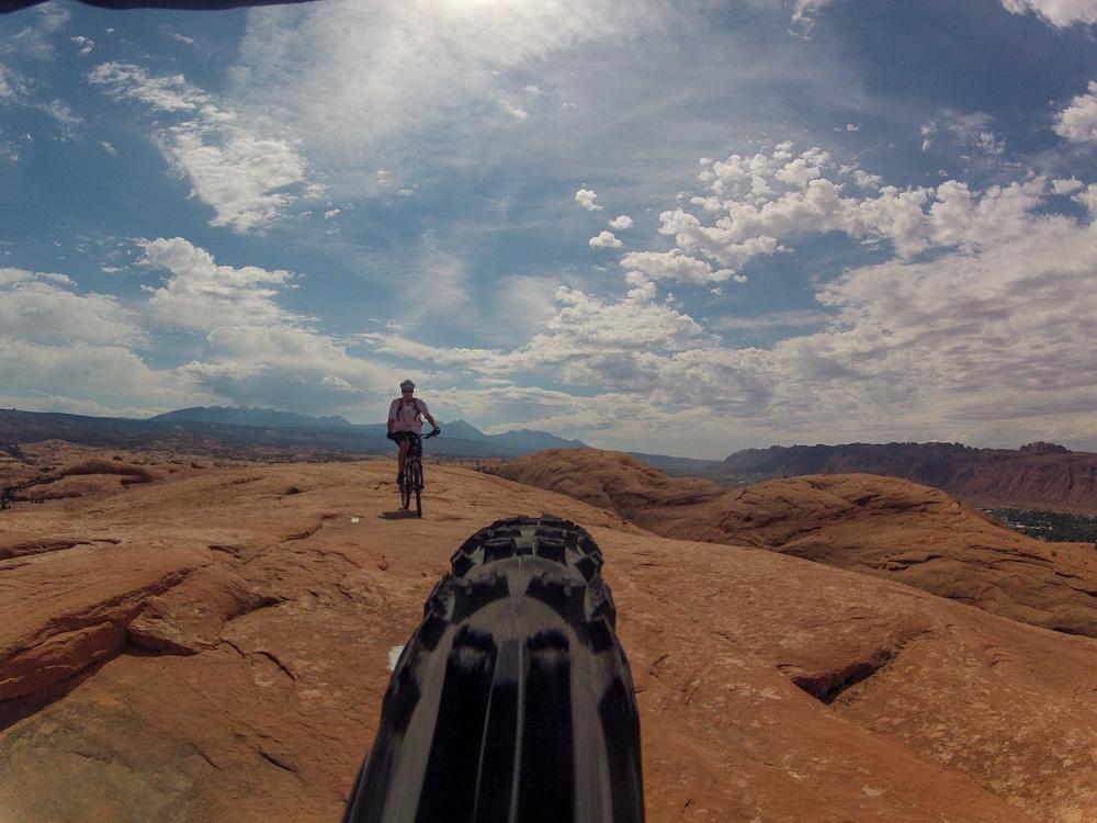 A mountain biker rides along a rocky trail under a blue sky with scattered clouds, with distant mountains visible in the background. The image is taken from a low angle, focusing on the bike tire in the foreground. Slickrock mountain bike trail.