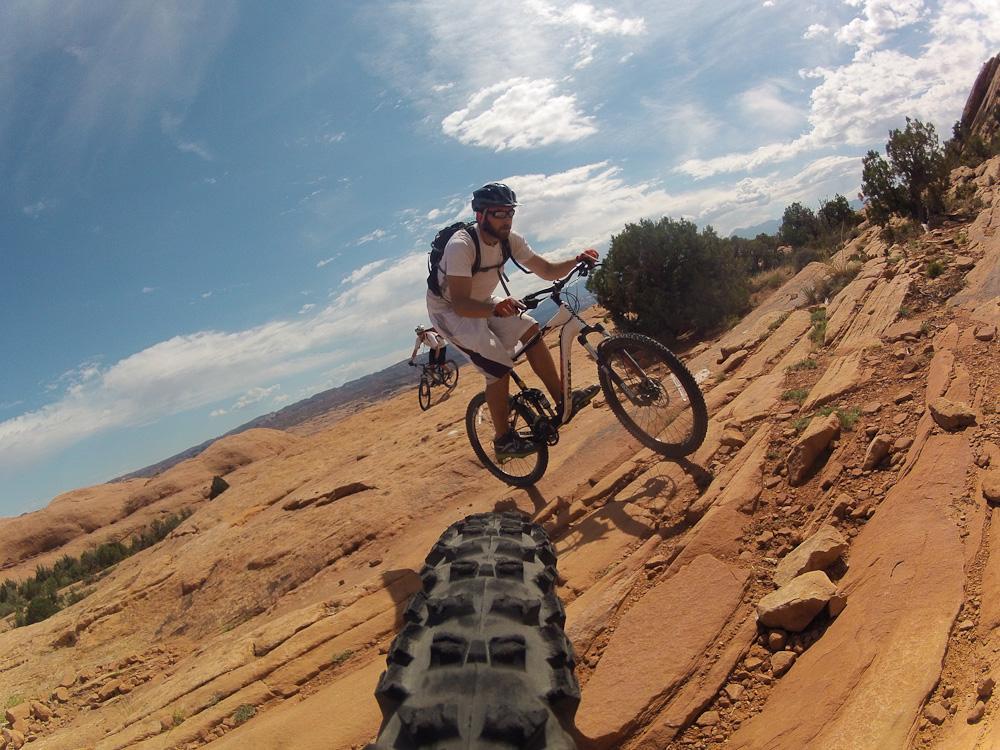 A person riding a mountain bike on a rocky trail, with a bright blue sky and clouds in the background. The image captures the dynamic movement of the cyclist as they navigate the terrain, with a close-up view of the bike's tire in the foreground. Slickrock mountain bike trail.