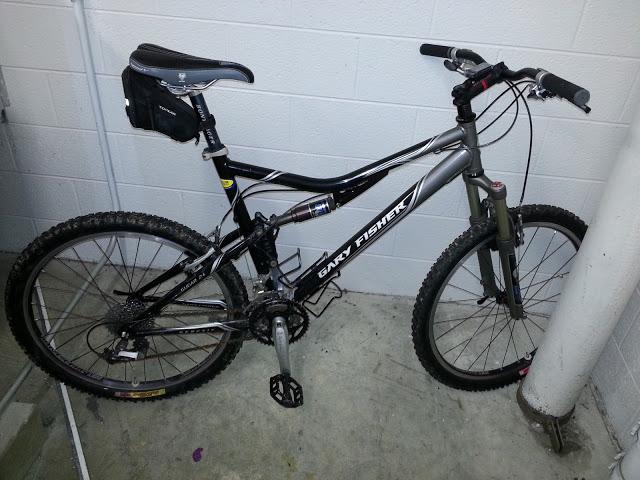 Gary Fisher Sugar 3+: Alt text: A black and silver Gary Fisher mountain bike leaning against a white wall, featuring thick tires and a small black bag attached to the frame. The bike is positioned on a concrete surface.