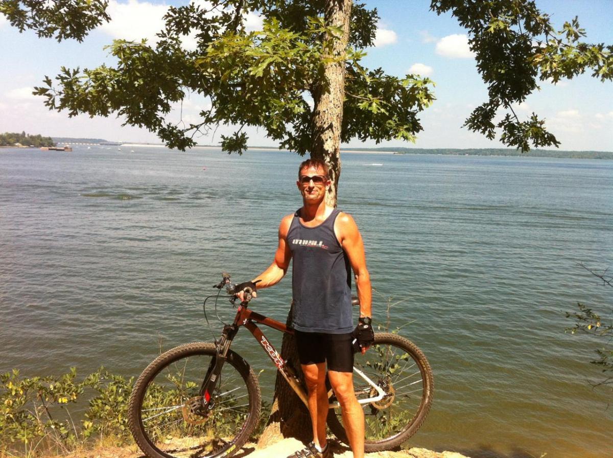 Kona Big Kahuna: A man stands next to a mountain bike by a calm lake, with a tree in the background. He is wearing a sleeveless shirt, shorts, and sunglasses, and he has a relaxed smile. The scene is sunny, showcasing a clear blue sky and greenery around the water's edge.