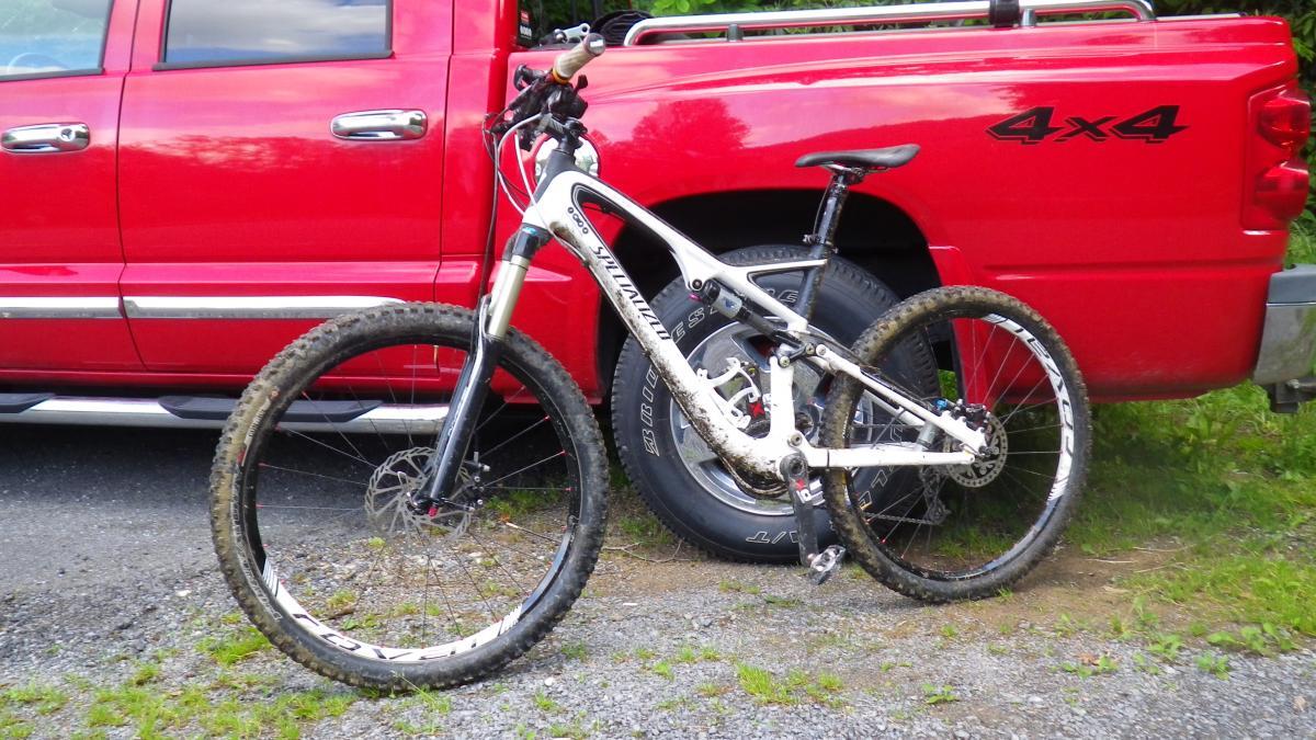 Specialized Stumpjumper FSR Pro Carbon: A mountain bike with a white and black frame leaning against a red pickup truck, parked on a gravel surface. The bike shows signs of use, with muddy tires, indicating it's been recently ridden on trails. The truck features a four-wheel-drive emblem on its rear.