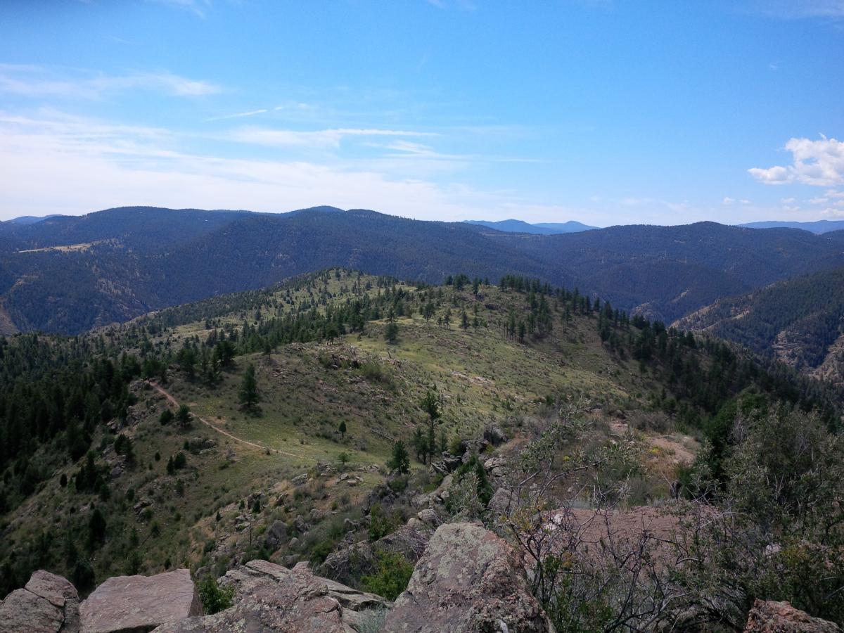A panoramic view of rolling green hills and distant mountains under a blue sky with scattered clouds, showcasing a natural landscape with patches of trees and rocky terrain. Centennial Cone Park mountain bike trail.