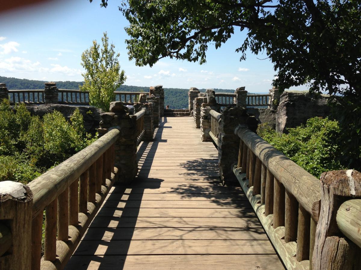 A wooden pathway leads toward a scenic overlook, bordered by stone railings and greenery on either side. In the distance, rolling hills and a blue sky are visible, indicating a peaceful natural setting. Coopers Rock mountain bike trail.