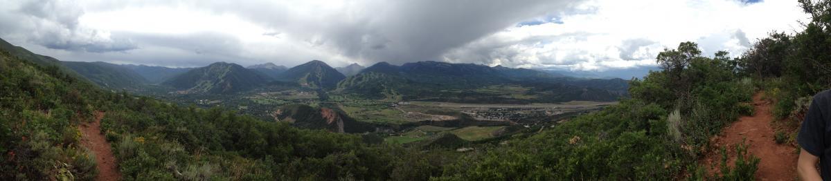A panoramic view of a mountainous landscape under a cloudy sky, with lush green vegetation in the foreground and a valley below. The mountains are rugged and prominent, while the valley features an open area that appears to be developed. The scene captures a tranquil, natural environment with varying shades of green and gray. Sunnyside Trail mountain bike trail.