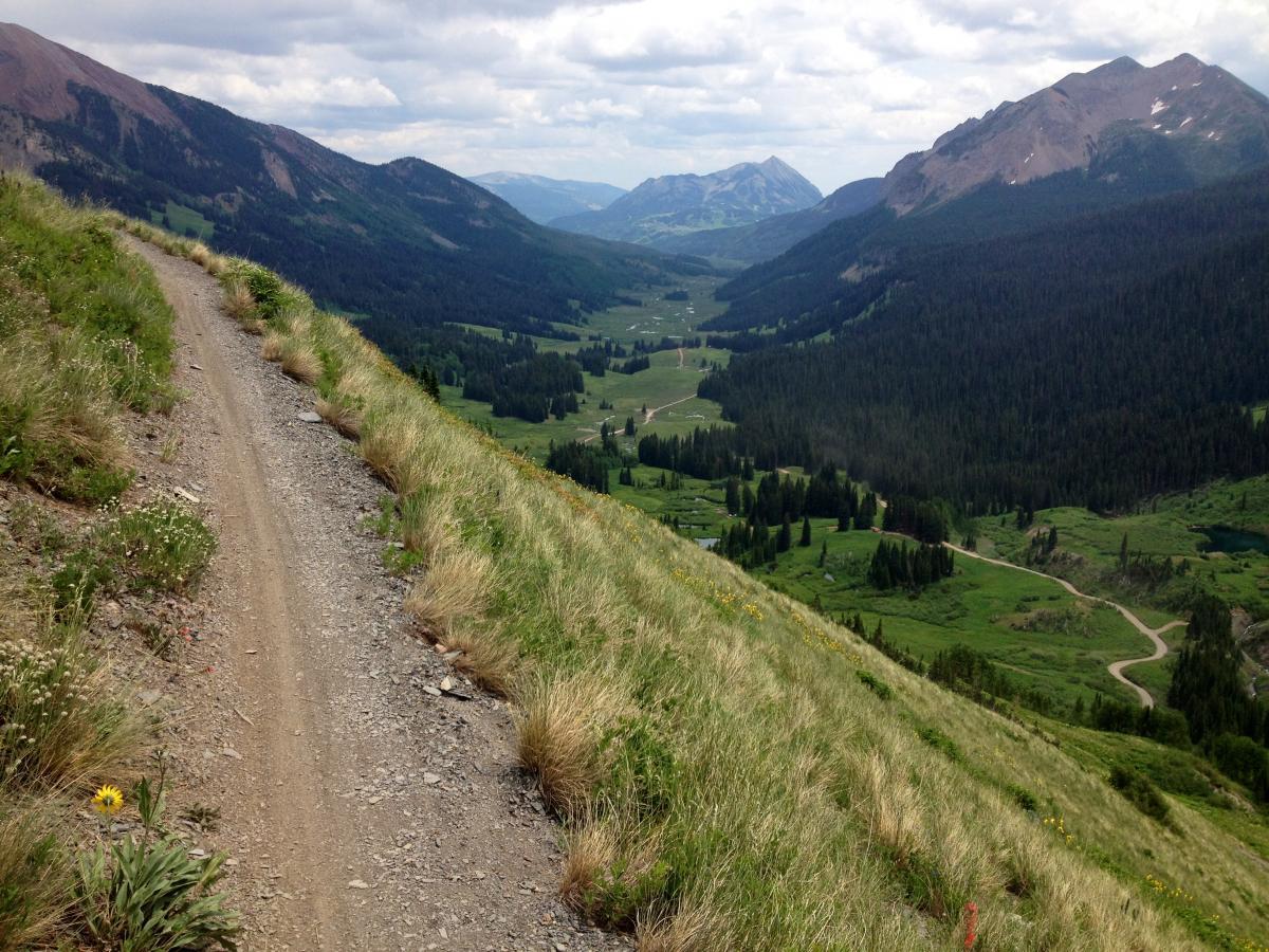 A winding dirt path bordered by tall grass leads down a hillside, with a panoramic view of lush green valleys and distant mountains under a partly cloudy sky. Trail 401 mountain bike trail.