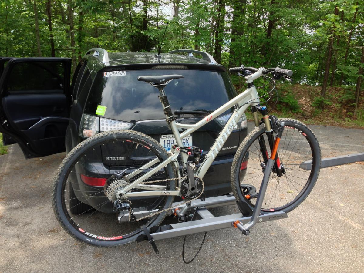 Niner Jet 9: An outdoor scene featuring a grey mountain bike mounted on a bike rack attached to a black Mitsubishi Outlander. The vehicle is parked on a paved surface with trees in the background, and the car's back door is open, suggesting a readiness for an outdoor adventure.