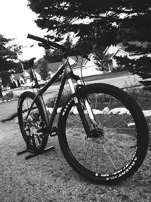 Kona Honzo: A black and white image of a mountain bike parked on a residential street. The bike is angled to show its frame, wheels, and tires, which feature brand markings. In the background, houses and trees are visible, hinting at a suburban setting.