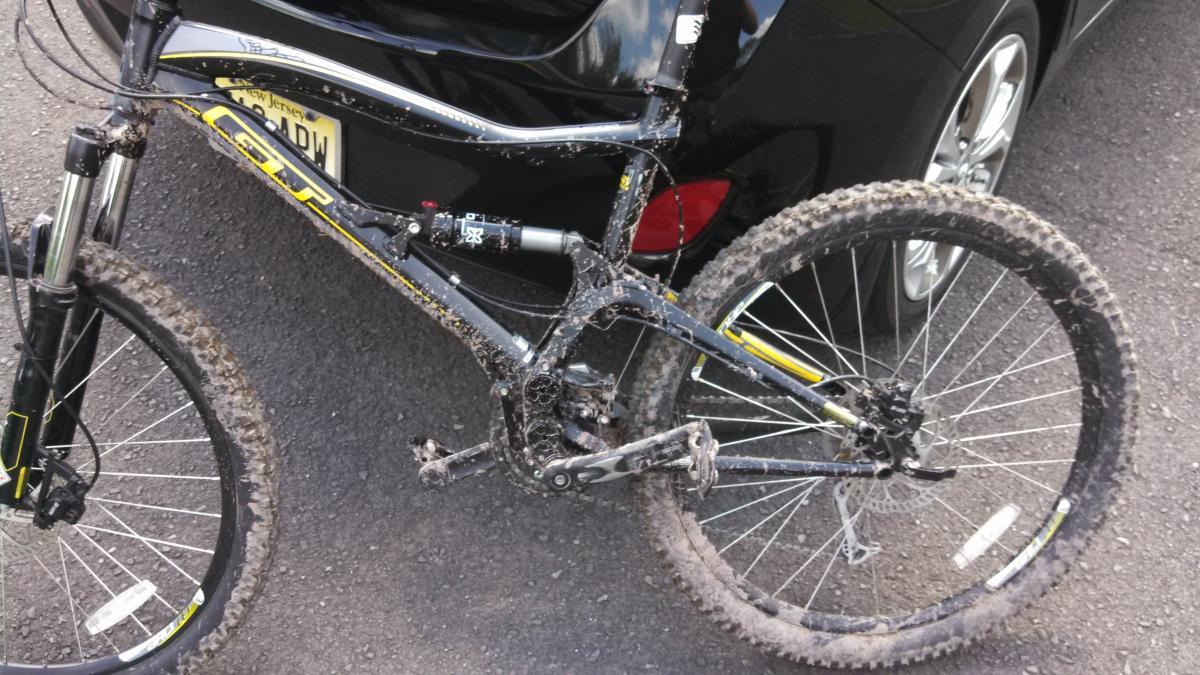GT Sensor: A close-up view of a muddy mountain bike parked next to a black car, showcasing its black and yellow frame, dirty tires, and suspension. The bike appears to have been recently used on a dirt trail.