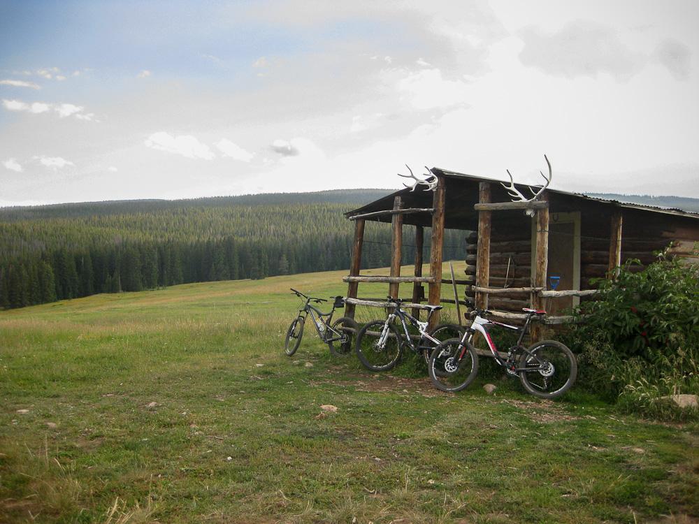 Bicycles parked near a rustic wooden shelter with antlers on the roof, set in a grassy landscape surrounded by dense evergreen trees under a cloudy sky. Meadow Mountain mountain bike trail.