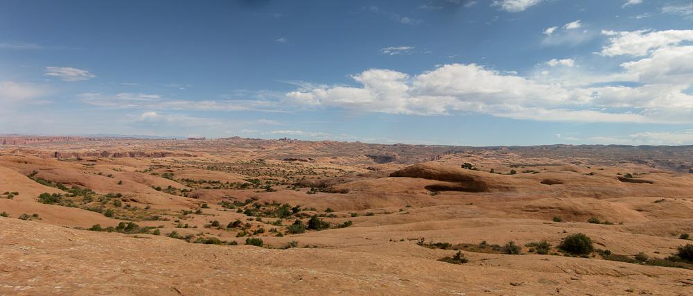 A panoramic view of a rugged, desert landscape featuring smooth, reddish-brown rock formations, scattered shrubs, and rolling hills. The sky is partly cloudy with varying shades of blue, suggesting a warm, sunny day. The expansive scene captures the natural beauty and arid conditions typical of desert environments. Slickrock mountain bike trail.