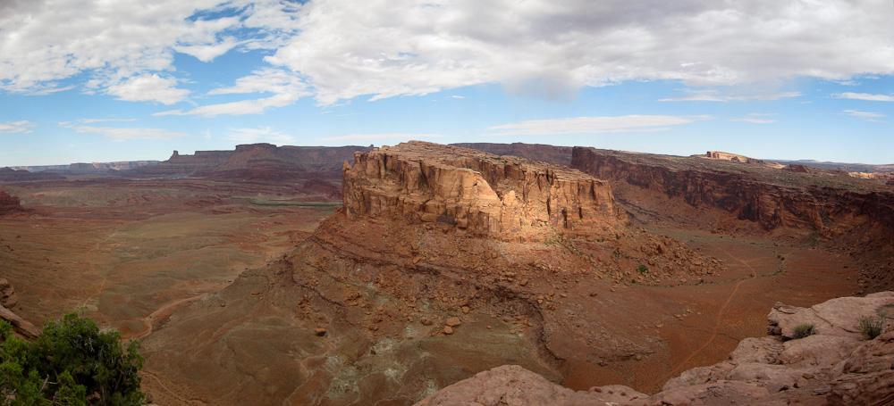 A panoramic view of a rocky landscape featuring towering cliffs and plateaus in a desert setting. The sky is partly cloudy, with blue patches visible, and the foreground includes sparse vegetation. The vast expanse of red and brown earth stretches into the distance, highlighting the natural beauty of the area. Amasa Back Trail mountain bike trail.