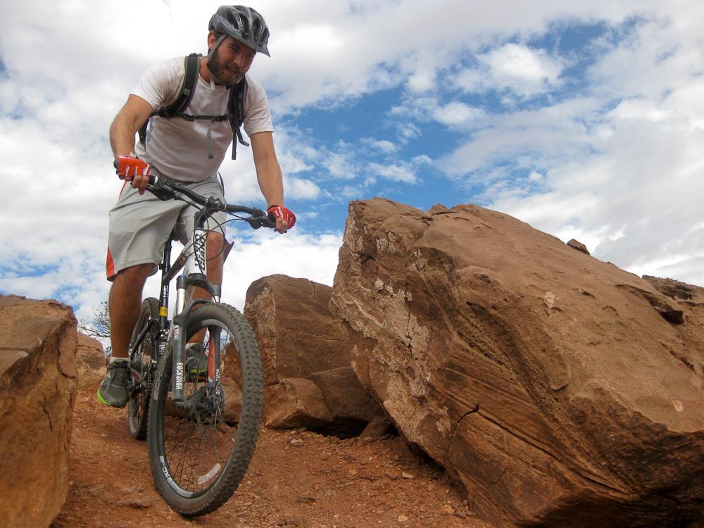 A man riding a mountain bike over rocky terrain, wearing a helmet and casual sportswear, with a blue sky and clouds in the background. Amasa Back Trail mountain bike trail.