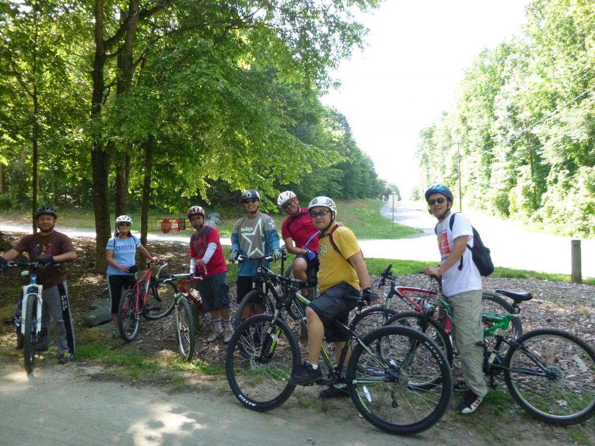 A group of seven people wearing helmets stands next to their bicycles on a sunny day in a wooded area. The background features a dirt path, trees, and a road sign indicating a stop ahead. They are smiling and appear ready to continue their biking adventure. Salem Lake mountain bike trail.