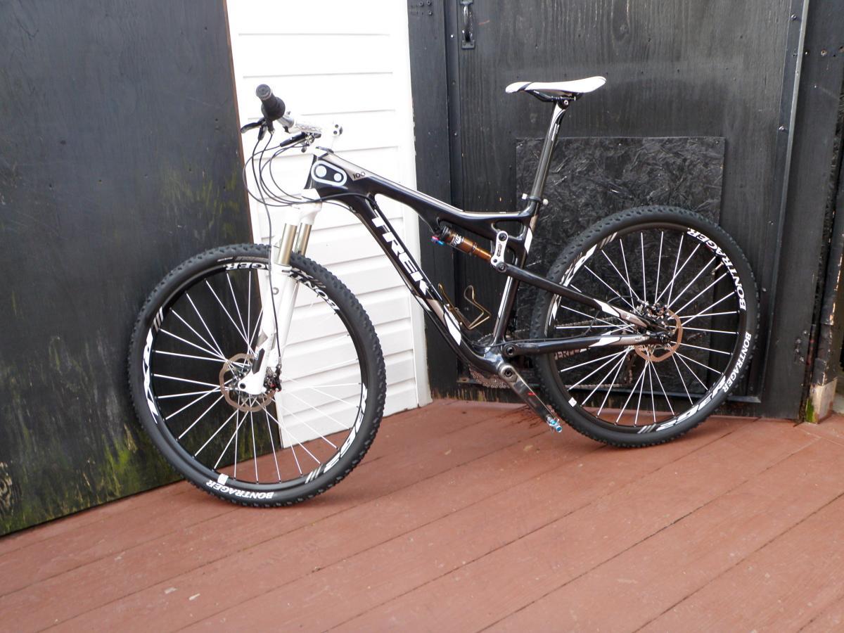 Trek Superfly Full Suspension: A black and white mountain bike leaning against a black wooden door. The bike features a lightweight frame, front suspension forks, and thick tires designed for off-road cycling. The background shows a wooden deck.