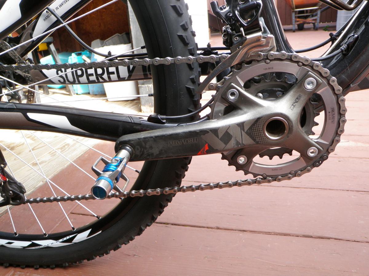 Trek Superfly Full Suspension: Close-up view of a mountain bike's drivetrain, featuring the crankset, chain, and rear wheel. The frame is partially visible with the word "SUPERFLY" printed on it, and the background shows a wooden deck and some blurred objects.
