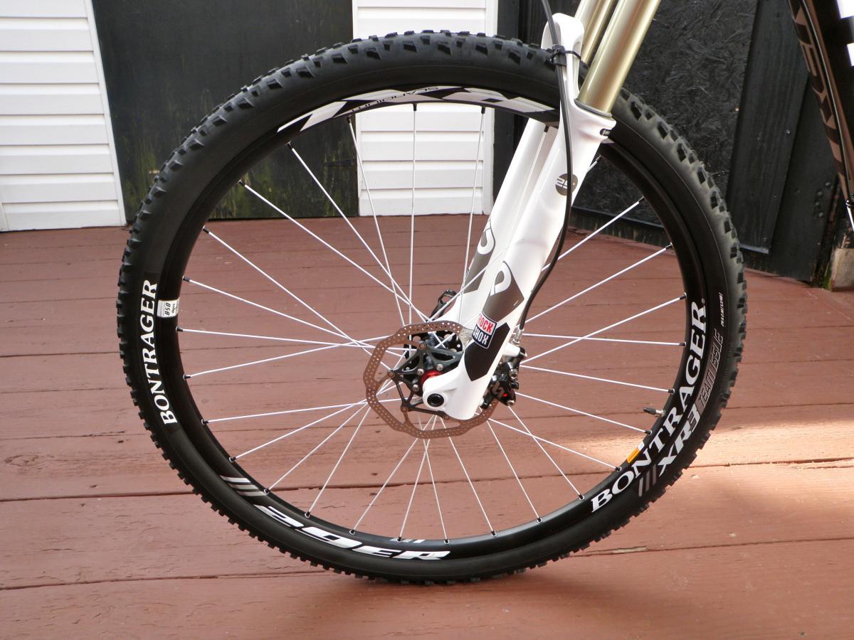 Trek Superfly Full Suspension: A close-up view of a mountain bike front wheel, featuring a Bontrager tire with rugged treads, mounted on a white bicycle frame with a suspension fork. The background shows a wooden deck and a partially visible building wall.