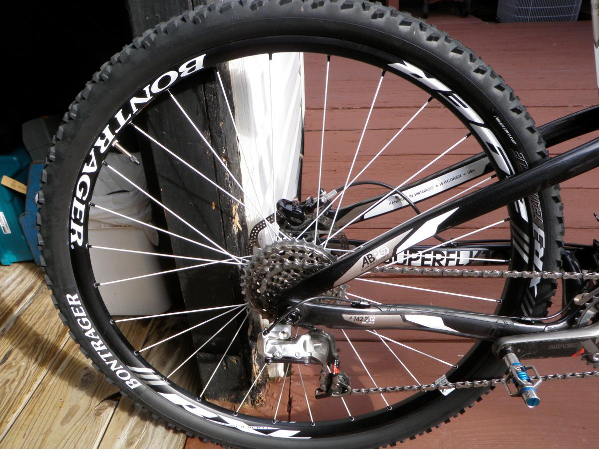 Trek Superfly Full Suspension: Close-up view of a mountain bike's rear wheel, featuring a Bontrager tire and cassette. The bike is positioned on a wooden deck with shadows cast and some equipment visible in the background.