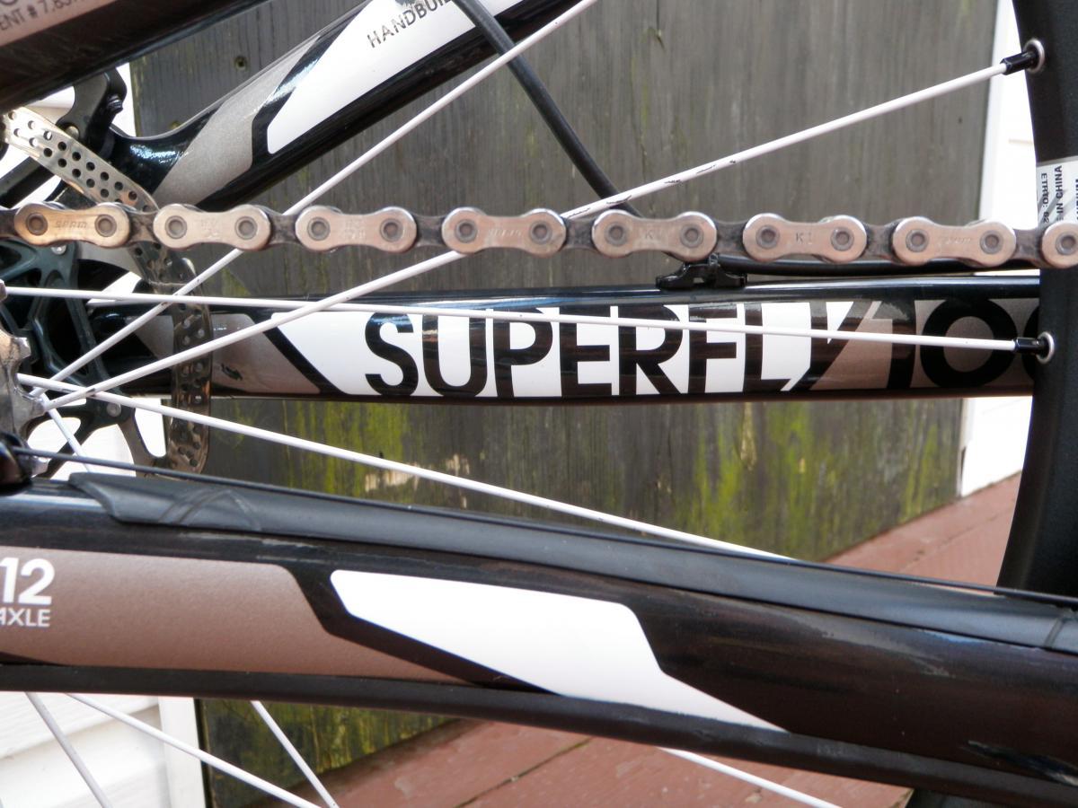 Trek Superfly Full Suspension: Close-up image of a bicycle's frame featuring the brand name "SUPERFLY" printed prominently, with a focus on the chain and rear derailleur. The background includes wooden planks and a rustic wall, adding context to the setting.