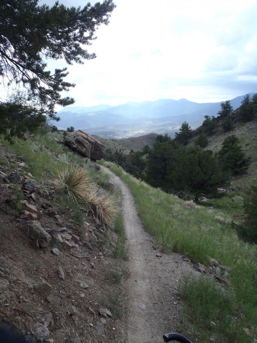 A winding dirt trail surrounded by greenery and rocky terrain, leading through a mountainous landscape. In the background, rolling hills and mountains are visible under a cloudy sky. A large boulder sits to the side of the trail, adding to the natural scenery. Lil Rattler mountain bike trail.