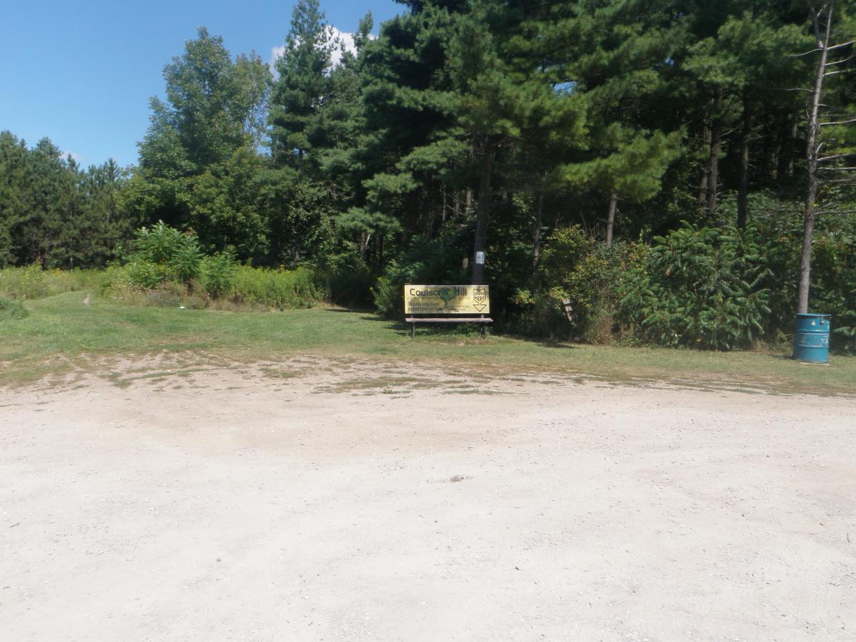 A gravel path leads to a wooded area, with a sign on a bench indicating the entrance to a park or nature reserve. Lush green trees and shrubs surround the area, creating a natural setting under a clear blue sky. A blue barrel is also visible in the scene. Coulson's Hill mountain bike trail.