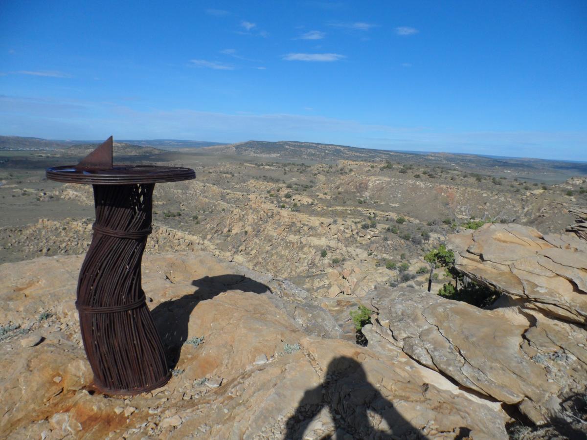 A twisted metal sculpture resembling a sundial stands on a rocky viewpoint, overlooking a vast, arid landscape with rolling hills and sparse vegetation under a clear blue sky. High Desert Trail System mountain bike trail.