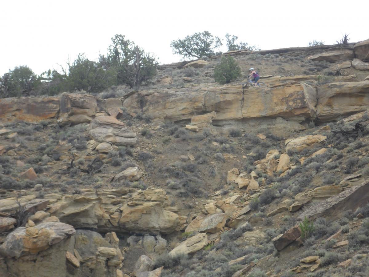 A mountain biker navigates a rocky, sloped terrain covered with sparse vegetation and rocky outcrops under a cloudy sky. High Desert Trail System mountain bike trail.