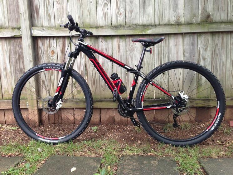 Trek Marlin: A black and red mountain bike leaning against a wooden fence, with a water bottle holder and rugged tires visible. The bike features two large wheels, a suspension fork, and disc brakes. The ground is covered with grass and stones.