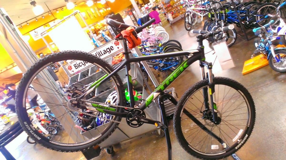 Trek Marlin: A black and green mountain bike displayed in a bicycle shop, with the front wheel lifted off the ground. The background features various other bicycles and cycling gear for sale, with a sign reading "Bicycle Village." Indoor lighting illuminates the store.