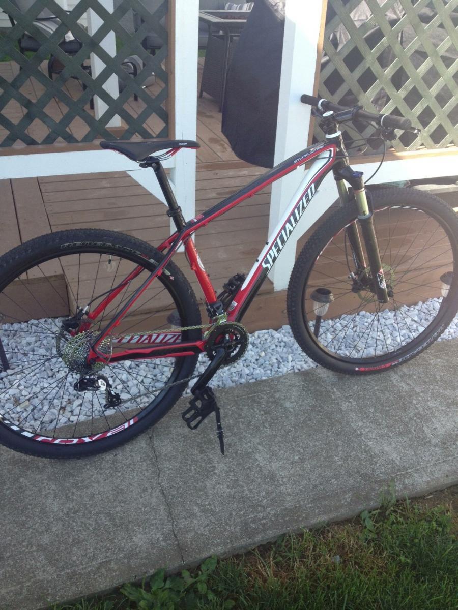 Specialized Stumpjumper Comp 29er: A red and black mountain bike leaning against a wooden deck, with a gravel area and grass in the foreground. The bike features thick tires, a sleek frame, and disc brakes. In the background, a patio area with outdoor furniture is visible.