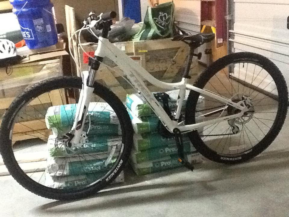 Trek Cali: A white mountain bike leaned against a stack of green and white bags in a garage setting. The bike features a front suspension fork, thick tires, and a visible drivetrain. Other items in the background include a blue bucket and various storage containers.