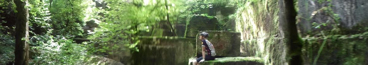 A person in a cycling helmet sits on a moss-covered rock in a lush, green forest, surrounded by dense foliage and rocky outcrops. Sunlight filters through the trees, creating a serene and tranquil atmosphere. Ijams Nature Centre mountain bike trail.