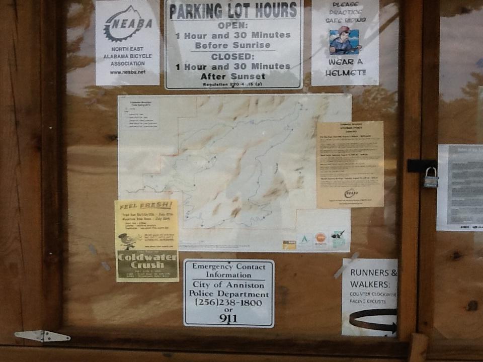 A wooden bulletin board displaying various signs and notices, including parking lot hours, a map, safety reminders about wearing helmets, and information about a local bicycle event. Emergency contact information for the City of Anniston Police Department is also visible. Coldwater Mountain mountain bike trail.