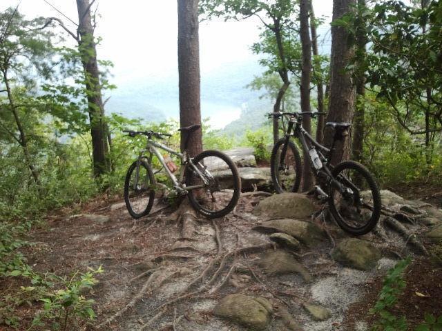 Two mountain bikes are parked on a rocky, root-covered clearing in a forest. In the background, trees frame a scenic view of a river and distant hills, suggesting a peaceful outdoor location. Raccoon Mountain Trail Network mountain bike trail.