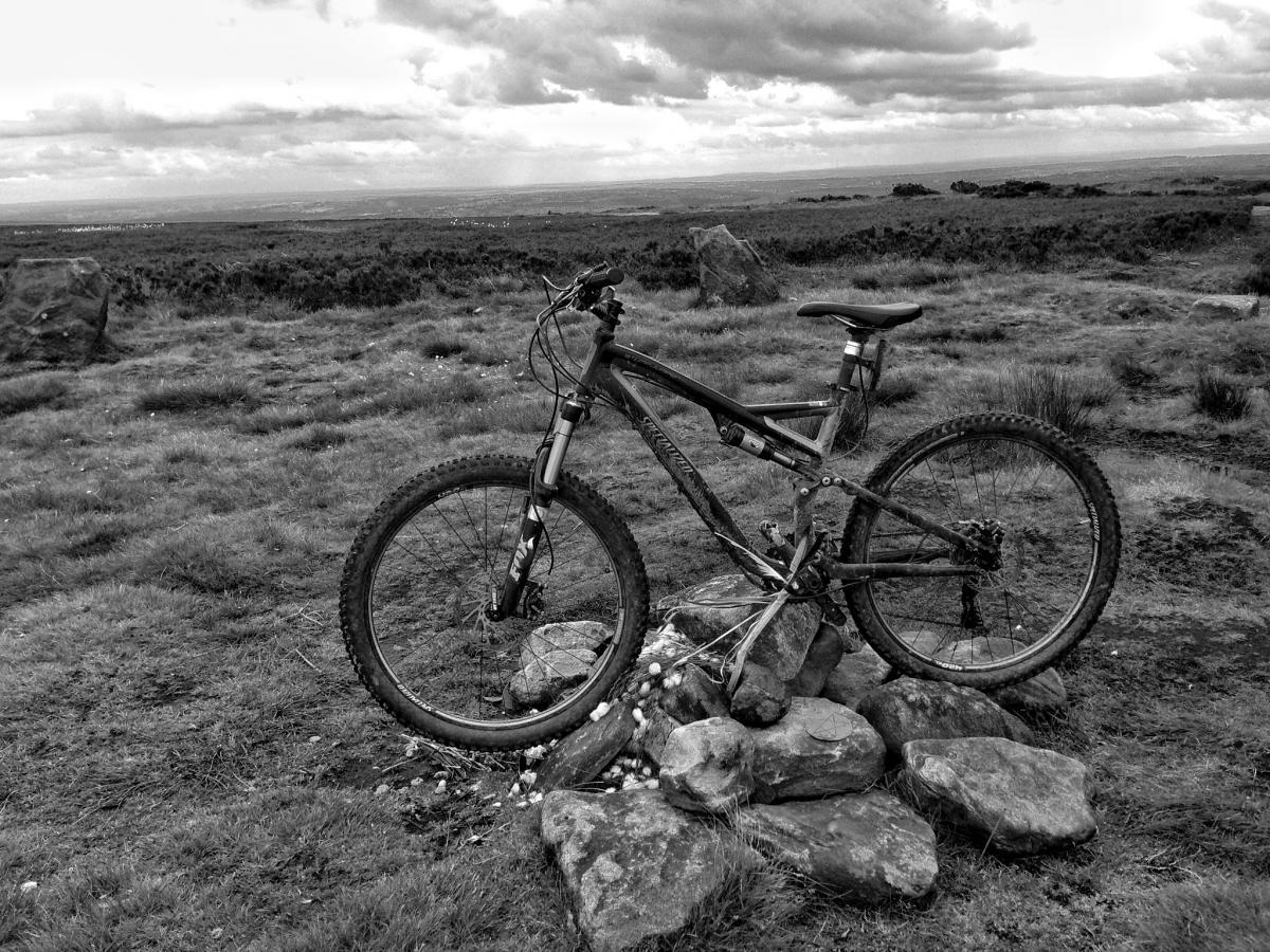Specialized Stumpjumper FSR: A mountain bike resting on a stack of rocks, set against a backdrop of open grassland and a cloudy sky, captured in black and white.