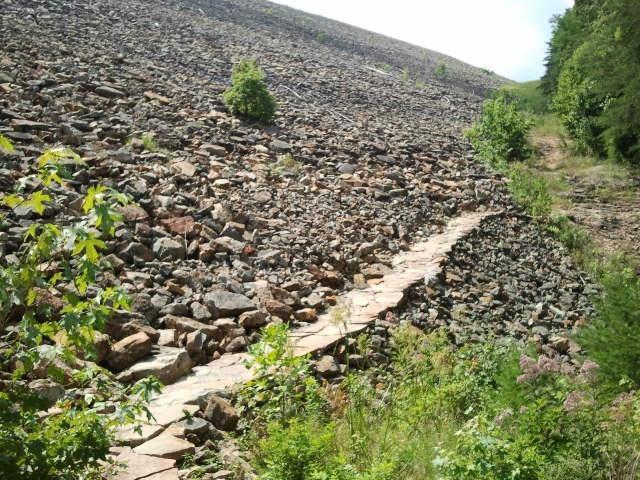 A winding stone path leads through a rocky terrain, surrounded by greenery and small patches of grass. The hillside in the background is steep and covered with various sizes of boulders. Sunlight illuminates the scene, creating a natural, tranquil atmosphere. Raccoon Mountain Trail Network mountain bike trail.