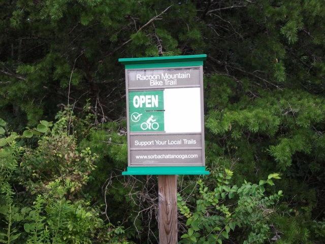 Sign for Raccoon Mountain Bike Trail indicating that the trail is open, with a bicycle symbol and a message encouraging support for local trails. The background features greenery typical of a forested area. Raccoon Mountain Trail Network mountain bike trail.
