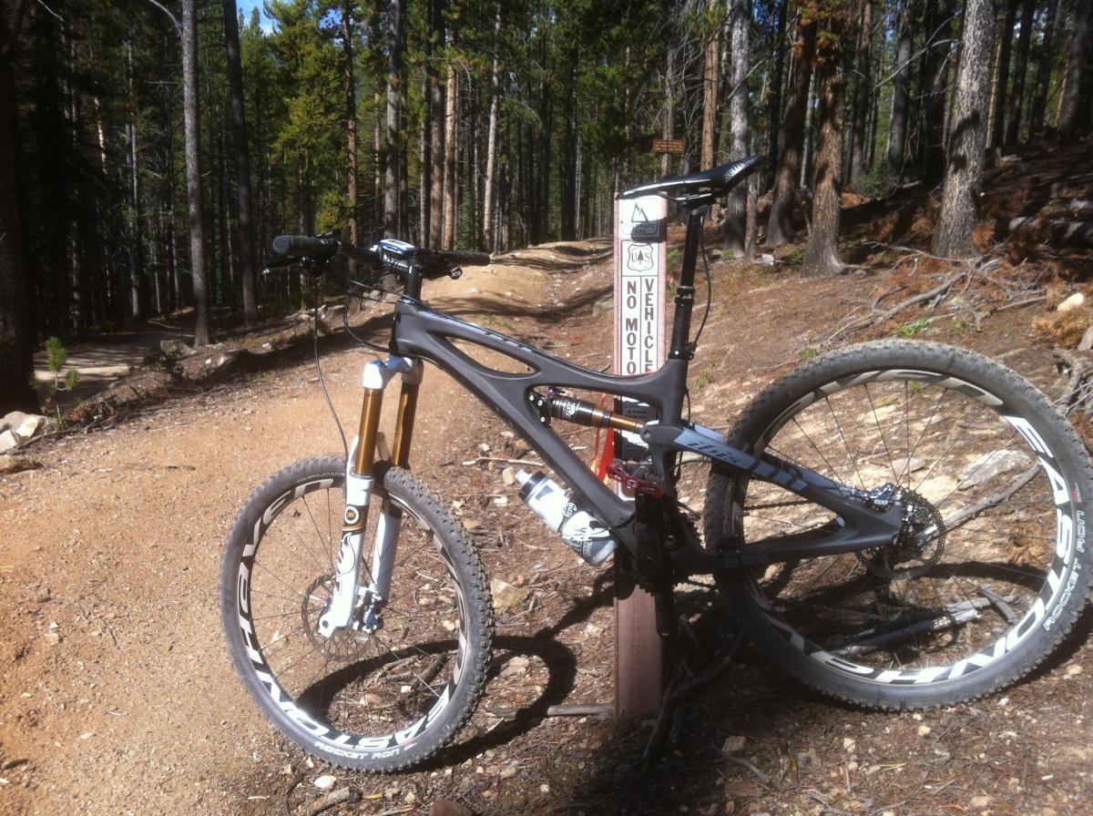 Ibis Mojo HD: A mountain bike parked beside a trail sign in a forest setting, with trees lining the path and a clear blue sky visible in the background. The bike features a sleek design with front suspension and disk brakes, resting on a dirt path. The sign warns that no motor vehicles are allowed on the trail.