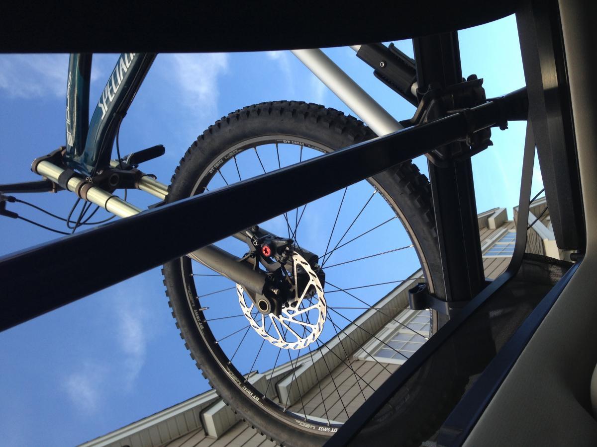 Specialized Enduro SL Expert: A view from inside a vehicle looking up at a mountain bike mounted on a roof rack, with blue sky and a building in the background. The bike's wheel and frame are prominently featured in the foreground.