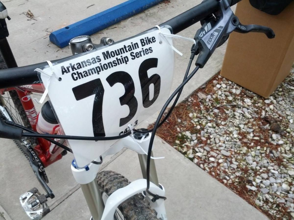 Jamis Nemesis: A close-up of a mountain bike's handlebars featuring a race bib labeled "736" for the Arkansas Mountain Bike Championship Series. The bike is positioned on a gravel surface with a blurred background of another bicycle and a cardboard box.