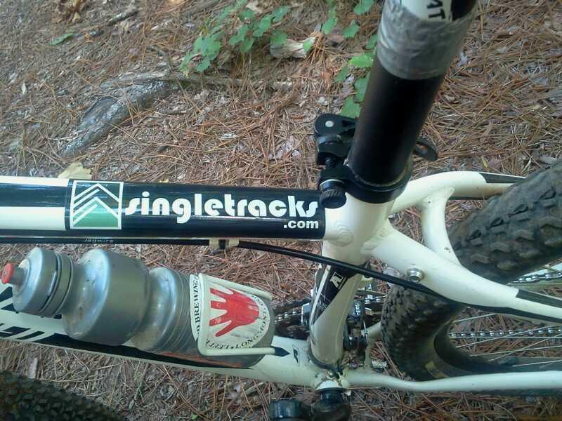 Close-up view of a mountain bike frame featuring a black and white branding sticker for "singletracks.com" on the top tube, along with a water bottle mounted on the frame. The background consists of pine needles and green foliage, suggesting an outdoor trail setting. Harbison State Forest mountain bike trail.