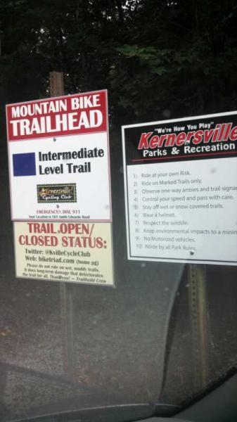 Signage at a mountain bike trailhead indicating an intermediate level trail. The sign provides safety guidelines and rules for trail usage, including reminders to ride at one's own risk and adhere to posted trail markers. Additional information about the trail's open or closed status is included at the bottom. Kernersville MTB park mountain bike trail.