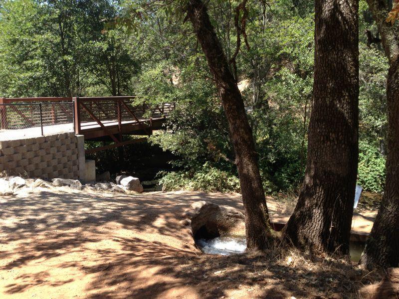A wooden pedestrian bridge spans a shallow stream surrounded by lush greenery. On the left, a stone wall leads down to the water, while a small rocky opening at the base allows water to flow out. Tall trees frame the scene, creating a tranquil, natural atmosphere. Hidden Falls Regional Park mountain bike trail.