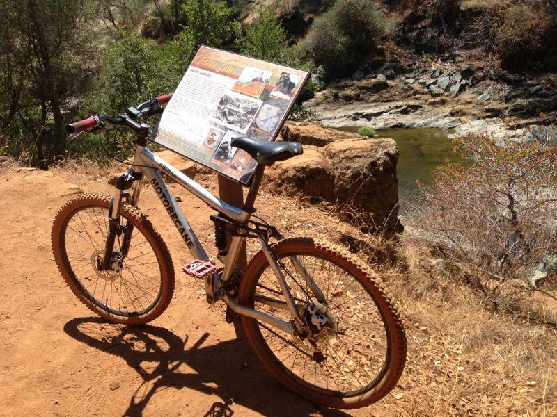 A mountain bike parked on a dirt trail beside a river, with a nearby informational sign featuring historical photos and text. Lush greenery surrounds the area, adding to the natural landscape. Hidden Falls Regional Park mountain bike trail.