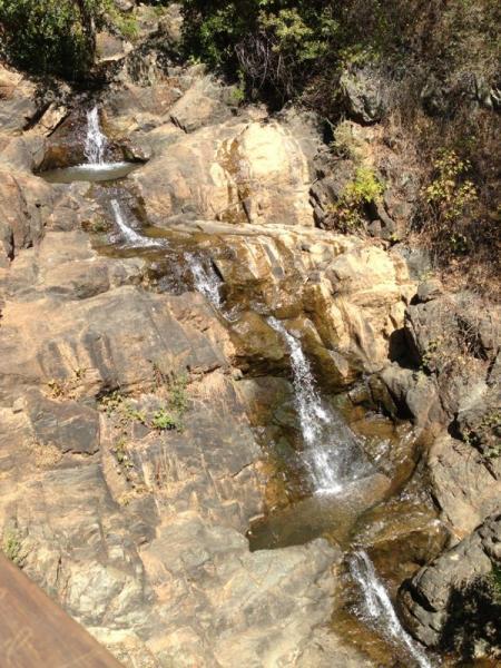 A cascading stream of water flows over a rocky terrain, surrounded by patches of greenery. The sunlight highlights the glistening water as it travels down the natural stone, creating small waterfalls amongst the rock formations. Hidden Falls Regional Park mountain bike trail.